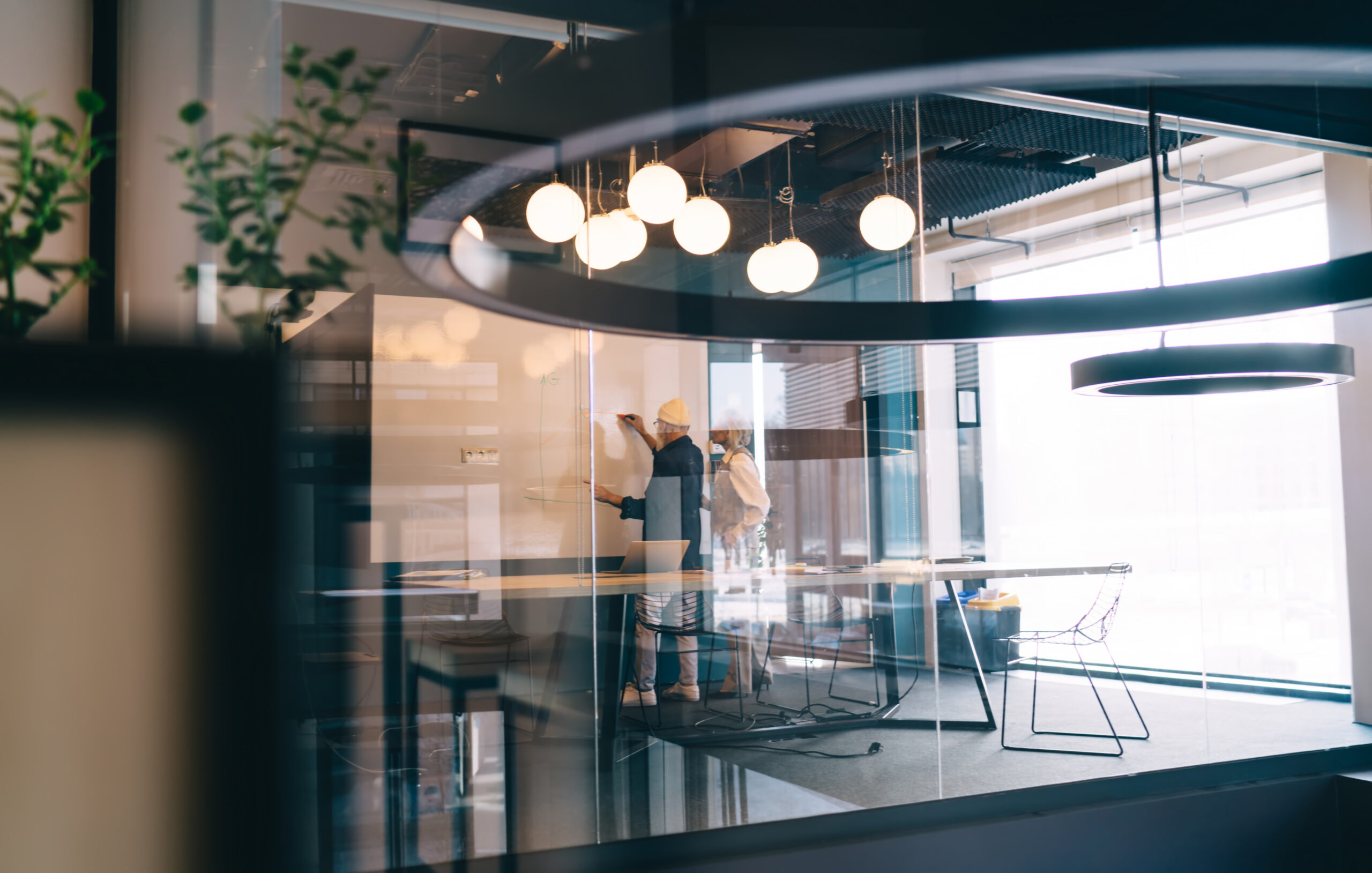 Through glass view of aged male writing on whiteboard while standing with aged female in meeting room with blurred glass wall with reflections of outside images and daylight